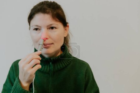 One beautiful Caucasian brunette girl with collected hair in a green sweater treats the right nasal passage with an apparatus with infrared light, sitting on a bed near a white wall, close-up side view with small copy space on the right.