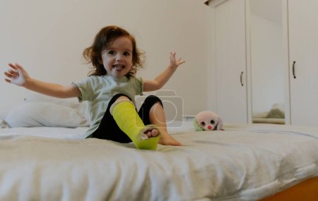 One beautiful little Caucasian girl with a green cast on her leg sits on the bed and cheerfully waves her arms with a happy smile on her face early in the morning in the room watching a cartoon on TV, side view close-up.