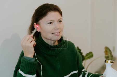 One beautiful Caucasian brunette girl with a smile on her face, gathered hair in a green sweater, treats her right ear with an infrared light device, sitting on a bed against a white wall, close-up