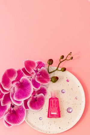 One small pink bottle of liquid soap lies below in a plate with one branch of an orchid flower and lilac glass stones on a soft pastel pink background with copy space on top, flat lay close-up.