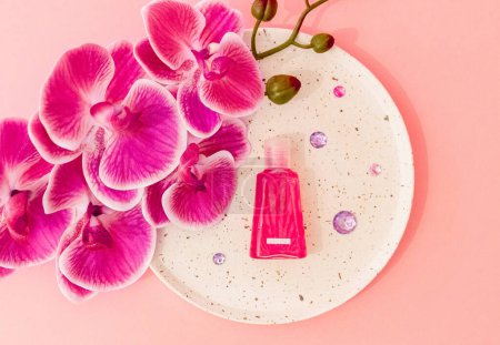 One small pink bottle of liquid soap lies in a plate with one branch of an orchid flower and lilac glass stones on a pastel pink background, flat lay close-up.