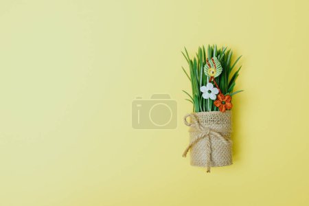 Photo for One beautiful homemade martisor of two flowers, a petal and a cheerful smiley face hangs on sprouted wheat in a jute pot lying on the right on a pastel yellow background with copy space on the left, flat lay close-up. - Royalty Free Image