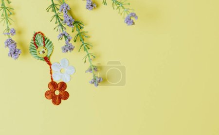Ein schönes hausgemachtes Martisor aus zwei Blumen, einem Blütenblatt und einem fröhlichen Smiley-Gesicht mit einem Strauß Frühlingsblumen liegt links vor pastellgelbem Hintergrund mit Kopierraum rechts, flache Lage in Nahaufnahme.