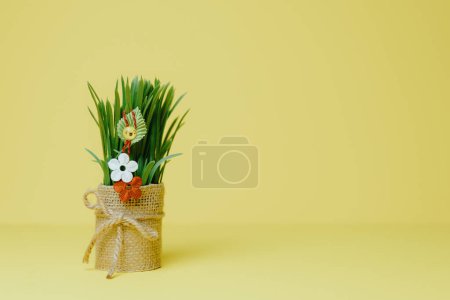 Photo for One beautiful homemade martisor of two flowers, a petal and a cheerful smiley face hanging on sprouted wheat in a jute pot stands on the left on a pastel yellow background with copy space on the right, side view close-up. - Royalty Free Image