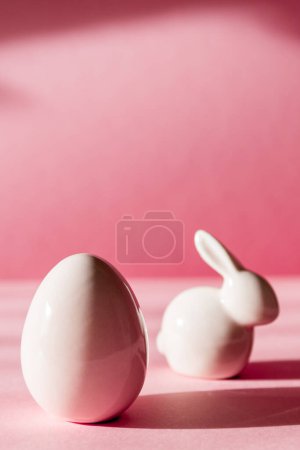 Porcelain figurines of Easter eggs and bunny stand on a pink background with shadows, close-up side view.
