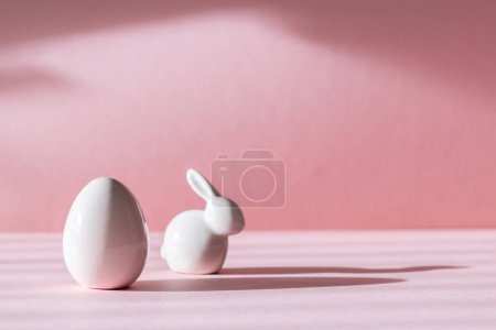 Porcelain figurines of Easter eggs and bunny stand on the left on a pink background with shadows and copy space on the right, side view close-up.