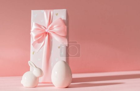 Porcelain figurines of Easter eggs and a big bunny gift box stand on the left on a pink background with shadows and copy space on the right, side view close-up.