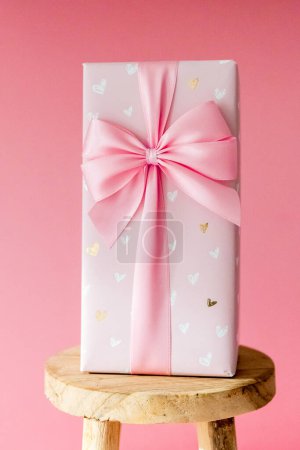 One large gift box with a bow stands on a small wooden stool on a pink background, close-up side view.