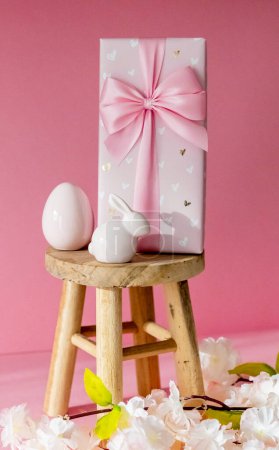 Porcelain Easter egg, bunny, large gift box with a bow stand on a small homemade decorative wooden stool on a pink background with a branch of apple tree blossom, side view close-up with selective focus.