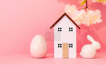 One wooden house with a porcelain Easter egg and bunny stands on the right on a pink background with small copy space on the left, side view close-up with selective focus.
