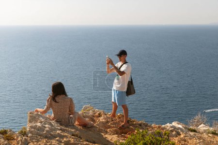 One cool guy in a cap takes a smartphone photo of a girl in a dress sitting on the top of a mountain and looking to the side against the backdrop of a blurry sea on a sunny summer day, close-up side view.