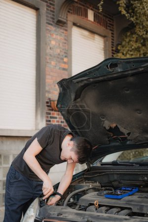 One young caucasian guy is unscrewing the burnt out bulb of the front right headlight of his car with a screwdriver while standing on a city street on a summer afternoon in the evening, close-up side