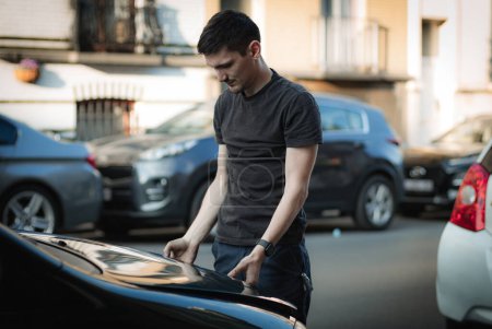 Ein junger kaukasischer Typ in dunkler Kleidung schließt die Motorhaube, nachdem er ein Auto repariert hat, das an einem Sommernachmittag am Abend halb seitlich auf einer Straße in der Stadt steht, Nahaufnahme von der Seite. Konzept für Ersatz
