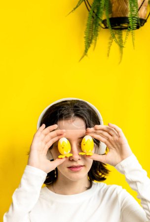 Téléchargez les photos : Portrait d'une jeune adolescente heureuse et blanche tenant avec ses doigts deux poulets décoratifs de Pâques au niveau des yeux, debout sur un fond jaune avec une fleur de palmier suspendue sur un - en image libre de droit