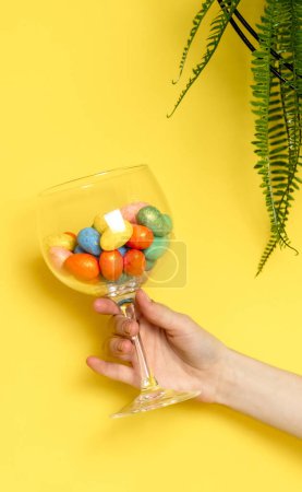 The hand of a young Caucasian unrecognizable girl holds a wine glass with Easter eggs and marble decorative eggs on a yellow background with a hanging palm flower, close-up side view.