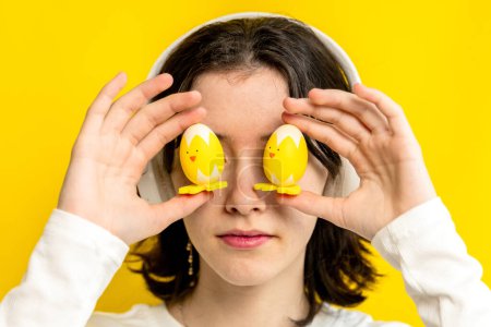 Téléchargez les photos : Portrait d'une jeune adolescente heureuse et blanche tenant avec ses doigts deux poulets décoratifs de Pâques au niveau des yeux, debout sur un fond jaune un jour de printemps dans la chambre, côté - en image libre de droit