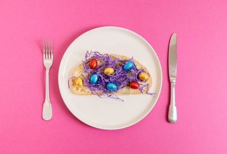 One sandwich with decorative lilac paper hay with chocolate Easter eggs in shiny multi-colored wrappers on a tray with a fork and knife lie in the center on a pink background, flat lay close-up