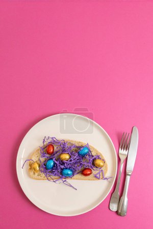 One sandwich with decorative lilac paper hay with chocolate Easter eggs in shiny multi-colored wrappers on a tray with a fork and knife lie from below on a pink background with copy space on top, flat
