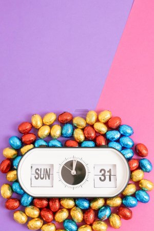 One alarm clock with the date and time of Easter 2024: Sunday March 31st with chocolate Easter eggs in colorful shiny wrappers lie from below on a lilac pink background with copy space on top, flat