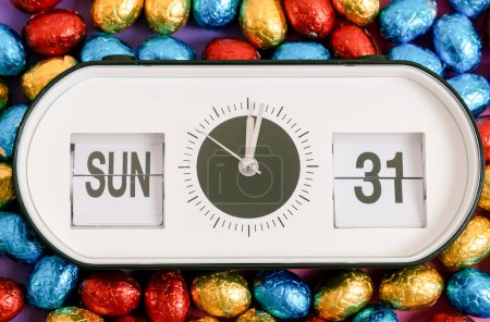 One alarm clock with date, time of Easter 2024: Sunday March 31st with chocolate Easter eggs in a colorful shiny wrapper on a lilac-pink background, flat lay very close-up. Concept of Easter candy