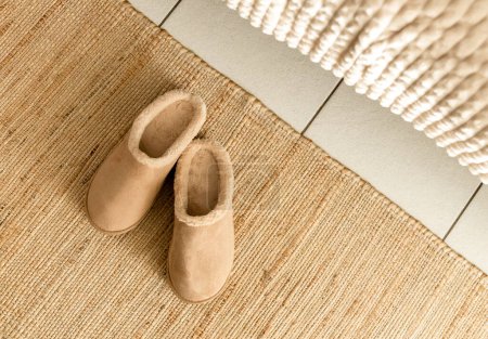 One pair of modern fashionable warm Uggs slippers lie on a woven jute rug by the bed in the bedroom, flat lay close-up. Fashionable shoes concept.