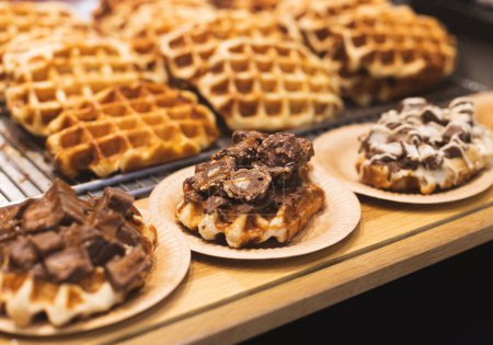 Appetitlich frische belgische Waffeln mit Schokoladenstücken in umweltfreundlichen Papptellern liegen auf einer hölzernen Backvitrine, Nahaufnahme mit Tiefenschärfe.