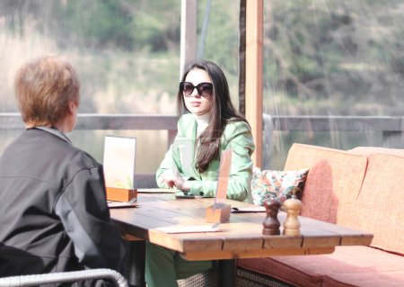Una joven hermosa psicóloga caucásica, una chica morena con un traje verde y gafas de sol, consulta a una mujer adulta sentada en la terraza de un café de agua en un soleado día de primavera, vista lateral de cerca.