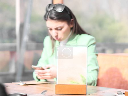 Téléchargez les photos : Un panneau vide pour un menu de restaurant se tient sur la table avec une fille triste et déçue floue avec un téléphone portable dans ses mains qui est assis sur la terrasse d'un café d'eau un jour de printemps ensoleillé - en image libre de droit