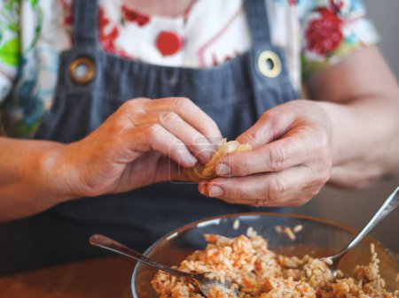 Hände einer Seniorin, die ein Blatt Sauerkraut mit Reisfleischfüllung umwickelt, sitzen an einem runden Tisch in der Küche, Seitenansicht in Großaufnahme. Das Konzept der Schritt-für-Schritt-Anleitung, Hausmannskost
