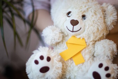 Little caucasian brunette female is holding in her hands a teddy bear with a yellow ribbon tie on a white wall background with a flower,close-up side view with depth of field. World childhood cancer