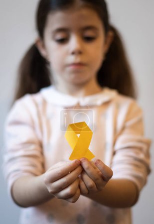 Beautiful little caucasian brunette girl with a sad face is holding out a yellow paper tape in her hands on a white background,close-up side view with depth of field. World childhood cancer day