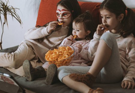 Three beautiful caucasian girls sisters are sitting on the sofa,eating corn rings and emotionally watching a movie on the tablet in the room in the evening,close-up side view.The concept of family