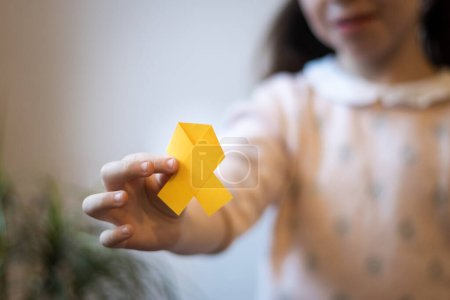 Beautiful little caucasian brunette female is holding a yellow paper tape in her hands on a white background,close-up side view with depth of field. World childhood cancer day concept.