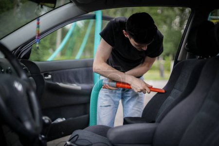 Un guapo joven caucásico morena chico sostiene un tubo con una boquilla en las manos y aspira el asiento en el coche, vista lateral de cerca.