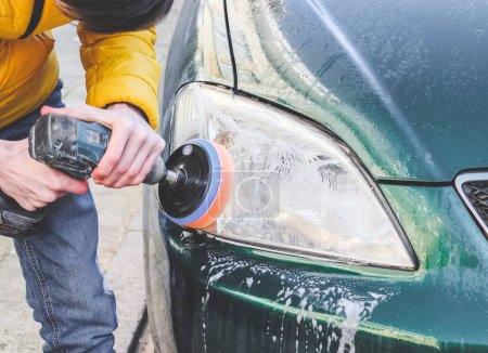 The hands of a young caucasian guy are drilling a drill with a disk with a sponge and polishing the headlight of his car with a detergent soap, close-up side view.The concept of a home car wash and