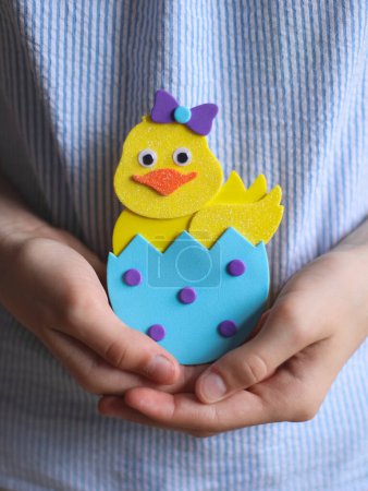 Hands of Caucasian teenage girl showing end result craft of yellow easter felt chicken with blue shell, orange beak, eyes, lilac bow and circles, holding in palms at belly level with blue striped