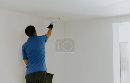 Un joven guapo hombre caucásico de cabello castaño con una camiseta azul se levanta de la espalda y pinta el techo al lado de la puerta con un rodillo de pintura blanca, vista lateral de cerca con enfoque selectivo