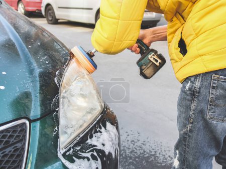 A young caucasian guy without a face in a yellow jacket is being drilled with a sponge disc and polishing the headlight of his car with a detergent soap on the city street in front of the house, close