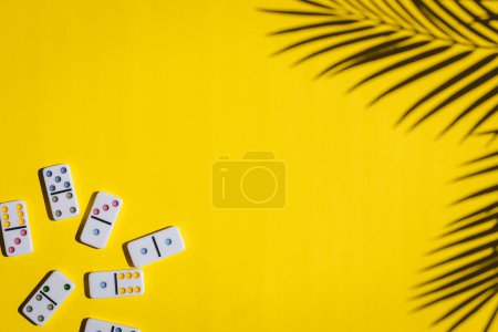 Weiße Dominosteine mit bunten Punkten liegen links auf gelbem Hintergrund mit dem Schatten eines Palmenzweiges und kopieren rechts, flach liegend, Nahaufnahme. Konzept Sommer Brettspiel.