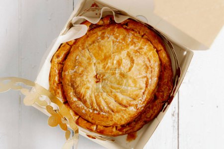 One royal galette in an open box with a golden crown lies on a white wooden table, flat lay close-up.