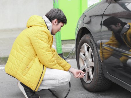 Young caucasian man in a yellow jacket and gray sweatpants, squatting, spins the bearing with his fingers into the wheel of a car for pumping air in a parking lot, side view with selective focus and