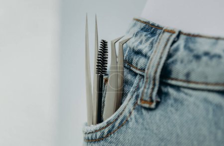 Zwei verschiedene Pinzetten und ein runder Pinsel für Wimpernverlängerungen ragen aus der rechten Tasche der Blue Jeans hervor, Nahaufnahme Seitenansicht mit kleinem Kopierraum links und Tiefenschärfe.
