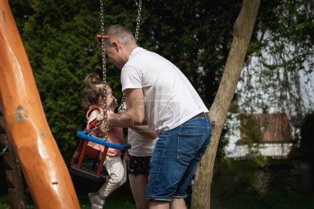 Un guapo joven caucásico padre toma a su pequeña niña llorando hija fuera del columpio en un día de verano en el parque en el patio de recreo, vista lateral.