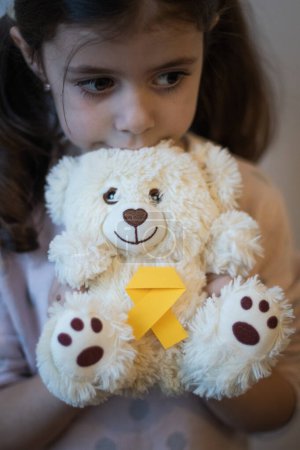 Beautiful little caucasian brunette female Holds in her hands holds a teddy bear with a yellow ribbon tie thoughtfully looking to the side on a white background,close-up side view with depth of field