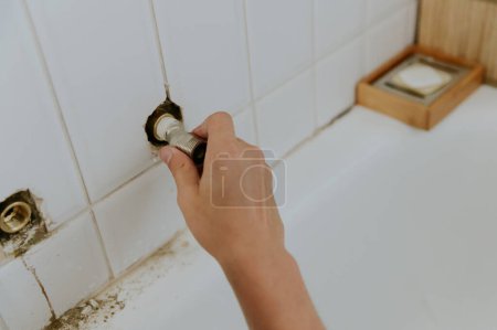 Téléchargez les photos : Un jeune homme caucasien méconnaissable sort un écrou de robinet d'un trou dans un mur de carrelage blanc tout en se tenant dans la salle de bain à la maison, vue latérale rapprochée.Étape par étape. - en image libre de droit