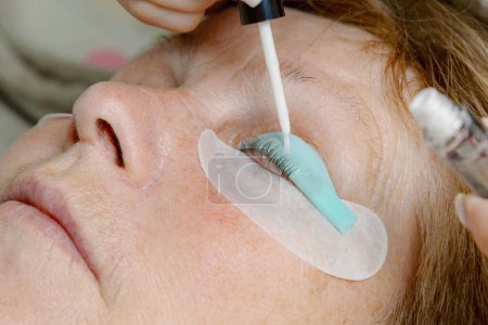 One Caucasian young unrecognizable girl cosmetologist applies transparent glue to the eyelashes of the left eye with a brush, pulling them onto a silicone stand of an elderly female client who is