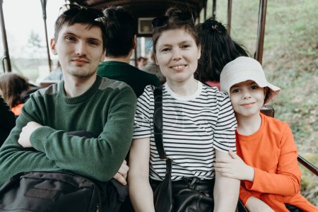 Portrait of one beautiful Caucasian family consisting of a young mother, an adult son and a little niece, looking with a smile at the camera and sitting in an open carriage of a tourist train