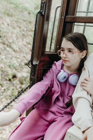 Ein Porträt eines schönen kaukasischen Mädchens mit Brille, einem helllila Jogginganzug, sitzt am Rand eines offenen hölzernen Retrowagens eines Touristenzuges, der durch ein Naturschutzgebiet fährt und blickt auf die