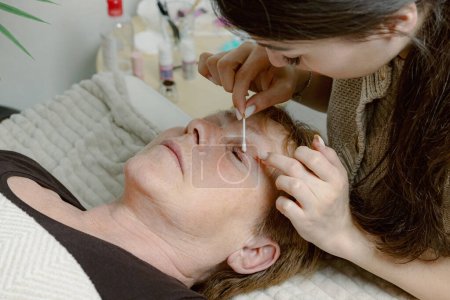 One Caucasian young beautiful brunette cosmetologist degreases the upper eyelid of the eye with a cotton swab to an elderly female client preparing them for the lamination procedure, which is lying on