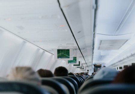 View of an electronic board with a flight path, inside a flying airplane with unrecognizable people sitting with their backs in chairs, close-up side view.